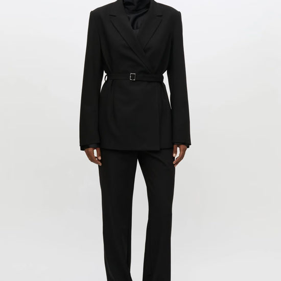 A model stands against a plain white studio background wearing a black tailored suit with a structured double-breasted blazer, a matching waist belt, a high folded collar, and matching trousers.