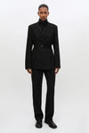 A model stands against a plain white studio background wearing a black tailored suit with a structured double-breasted blazer, a matching waist belt, a high folded collar, and matching trousers.