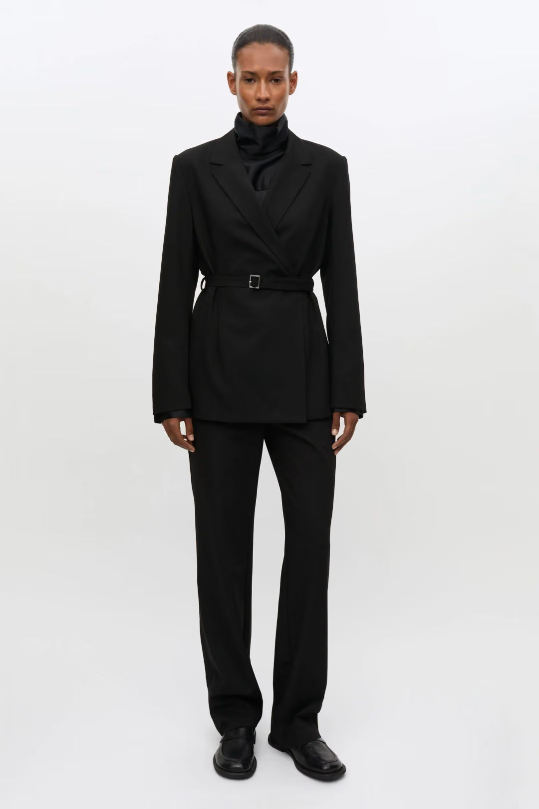 A model stands against a plain white studio background wearing a black tailored suit with a structured double-breasted blazer, a matching waist belt, a high folded collar, and matching trousers.