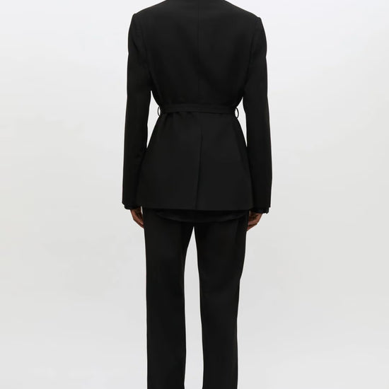 A model viewed from the back wearing a black tailored suit with a structured blazer and matching trousers, photographed against a clean white studio background.