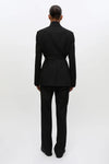 A model viewed from the back wearing a black tailored suit with a structured blazer and matching trousers, photographed against a clean white studio background.