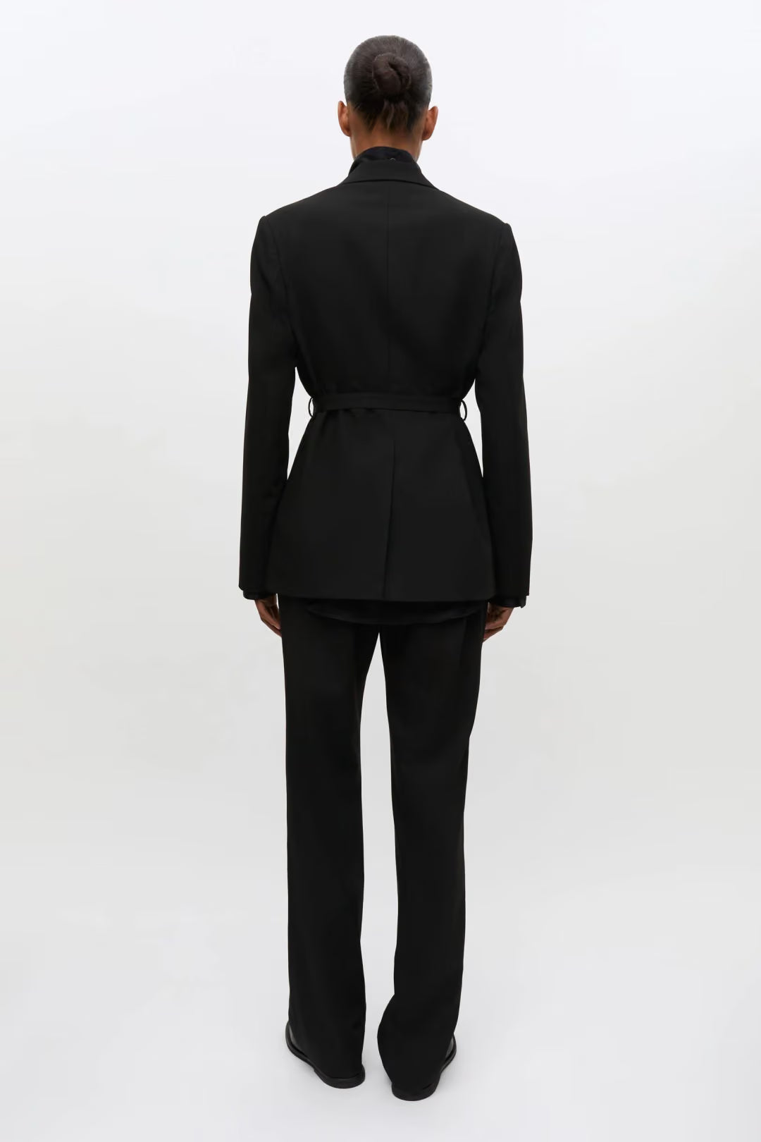 A model viewed from the back wearing a black tailored suit with a structured blazer and matching trousers, photographed against a clean white studio background.