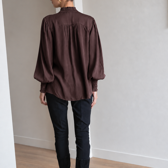 A woman viewed from behind wearing a dark brown long-sleeve blouse with gathered shoulders, black pants, and black knee-high boots, standing in a minimalist room with light gray walls and a wood floor.