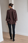 A woman viewed from behind wearing a dark brown long-sleeve blouse with gathered shoulders, black pants, and black knee-high boots, standing in a minimalist room with light gray walls and a wood floor.