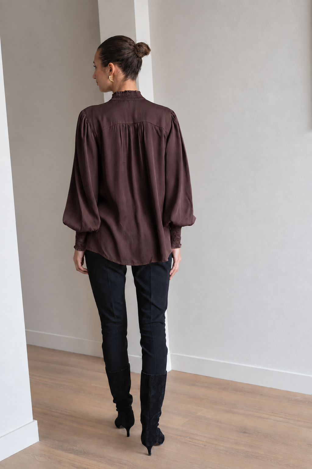 A woman viewed from behind wearing a dark brown long-sleeve blouse with gathered shoulders, black pants, and black knee-high boots, standing in a minimalist room with light gray walls and a wood floor.