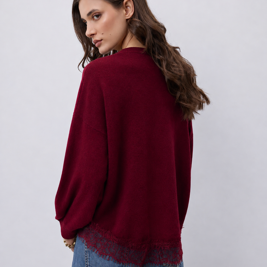 A woman wearing a deep burgundy knit sweater with a lace hem, paired with blue jeans, photographed from the back against a light gray studio background.
