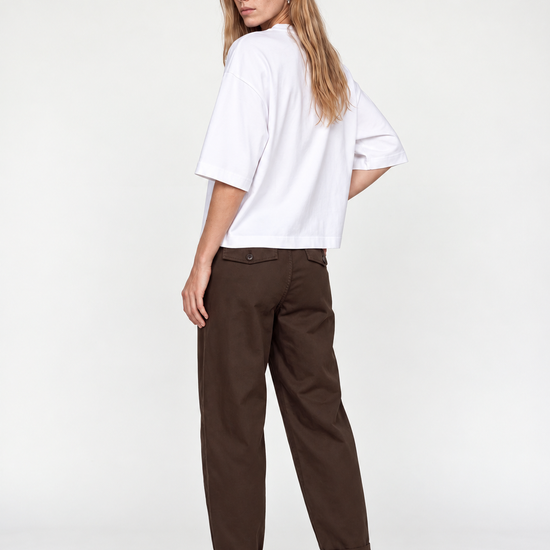 A woman modeling a white short-sleeve top and dark brown trousers, shown from a back-three-quarter angle against a plain white studio background.