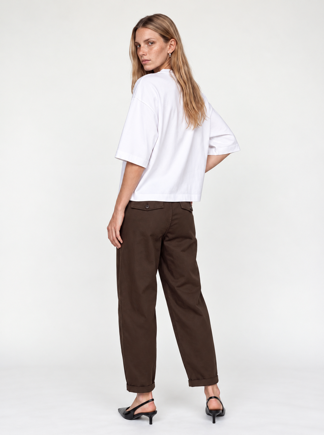 A woman modeling a white short-sleeve top and dark brown trousers, shown from a back-three-quarter angle against a plain white studio background.