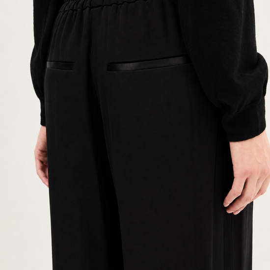 Close-up of a person wearing black wide-leg trousers with a gathered elastic waistband and black satin-like trim details, paired with a black long-sleeve top against a plain light background.