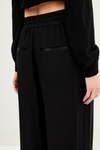 Close-up of a person wearing black wide-leg trousers with a gathered elastic waistband and black satin-like trim details, paired with a black long-sleeve top against a plain light background.