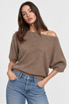 A woman wearing a brown off-the-shoulder knit sweater and blue jeans, photographed against a plain light gray studio background.