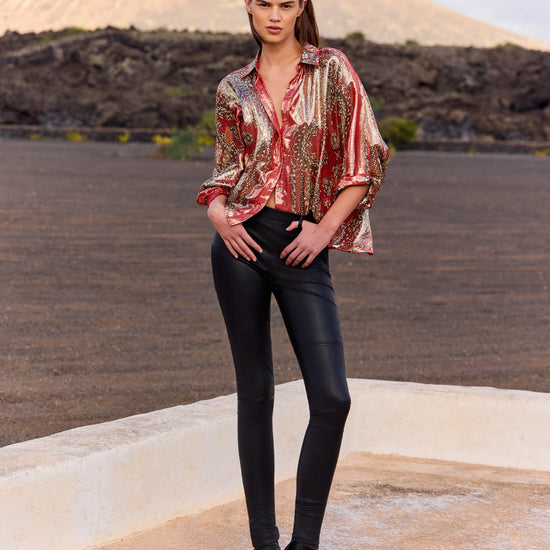 Woman wearing a patterned blouse and black pants standing on a rooftop with a desert landscape in the background.