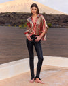 Woman wearing a patterned blouse and black pants standing on a rooftop with a desert landscape in the background.