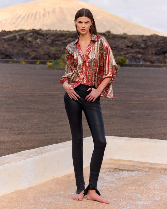 Woman wearing a patterned blouse and black pants standing on a rooftop with a desert landscape in the background.