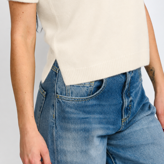 Close-up of a person wearing a cream short-sleeve knit top and blue denim jeans against a light gray studio background.