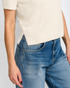 Close-up of a person wearing a cream short-sleeve knit top and blue denim jeans against a light gray studio background.