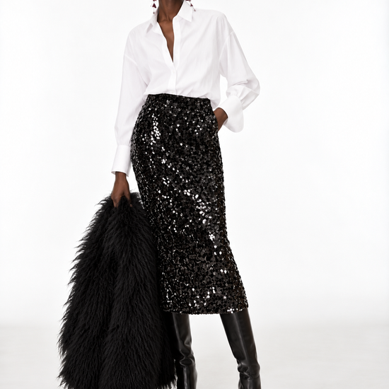 A model stands against a white studio background wearing a white button-up shirt, a black sequined midi skirt, black knee-high boots, and holding a large black feather boa.