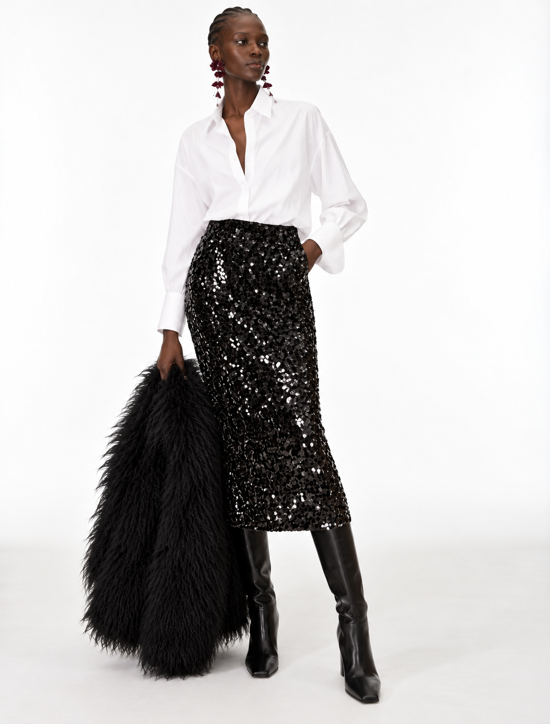 A model stands against a white studio background wearing a white button-up shirt, a black sequined midi skirt, black knee-high boots, and holding a large black feather boa.