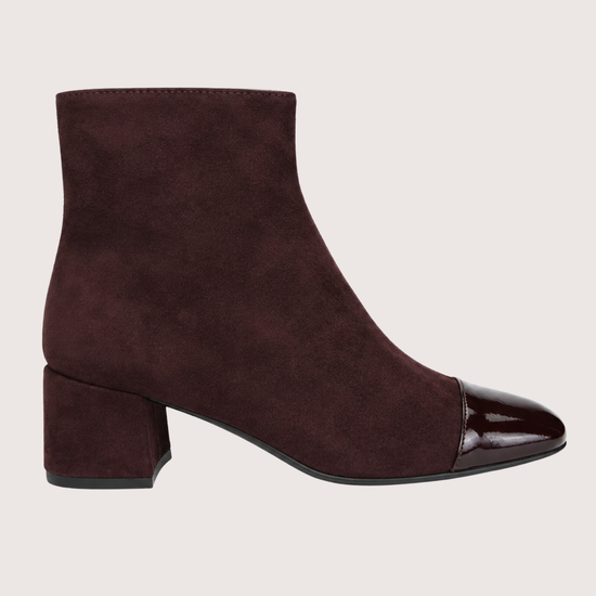 A dark burgundy suede ankle boot with a glossy patent leather cap toe and a low block heel, shown in profile against a light beige background.