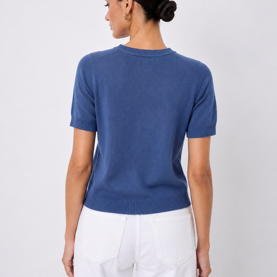 A woman shown from the back wearing a short-sleeve crewneck knit sweater in a medium blue color, paired with white pants, against a light gray studio background.