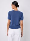 A woman shown from the back wearing a short-sleeve crewneck knit sweater in a medium blue color, paired with white pants, against a light gray studio background.