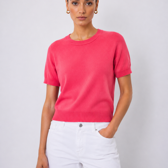 A woman modeling a short-sleeve crewneck knit sweater in a vibrant pinkish-red color, paired with white pants, against a light gray studio background.
