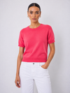 A woman modeling a short-sleeve crewneck knit sweater in a vibrant pinkish-red color, paired with white pants, against a light gray studio background.