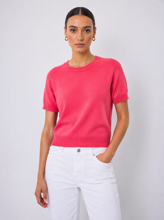 A woman modeling a short-sleeve crewneck knit sweater in a vibrant pinkish-red color, paired with white pants, against a light gray studio background.
