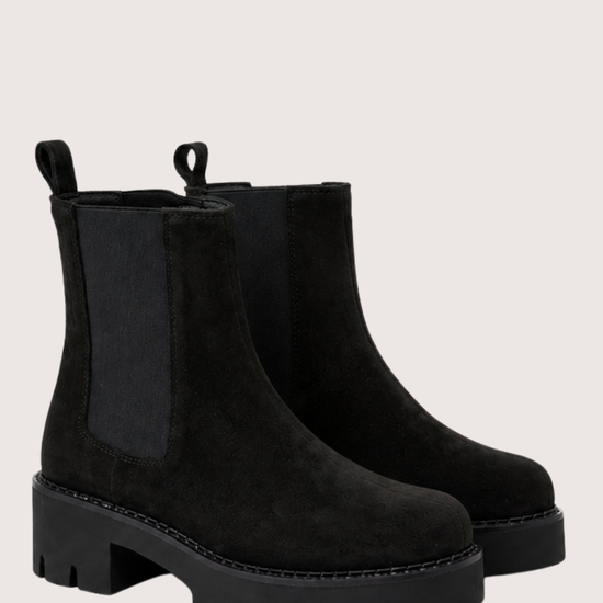 A pair of black suede Chelsea-style ankle boots with black elastic side panels and chunky lug soles, shown on a light beige studio background.