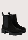 A pair of black suede Chelsea-style ankle boots with black elastic side panels and chunky lug soles, shown on a light beige studio background.
