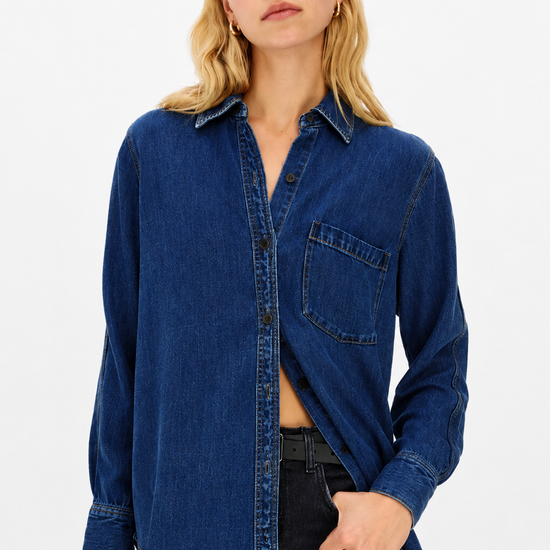 A woman wearing a dark blue denim button-up shirt with a chest pocket, paired with dark jeans, against a plain white studio background.