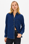 A woman wearing a dark blue denim button-up shirt with a chest pocket, paired with dark jeans, against a plain white studio background.