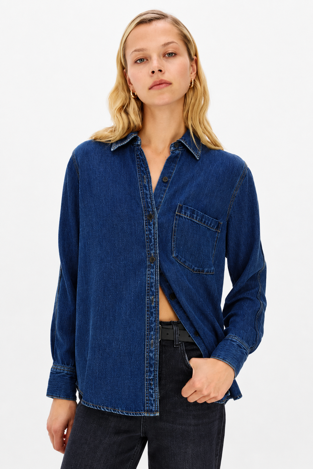 A woman wearing a dark blue denim button-up shirt with a chest pocket, paired with dark jeans, against a plain white studio background.