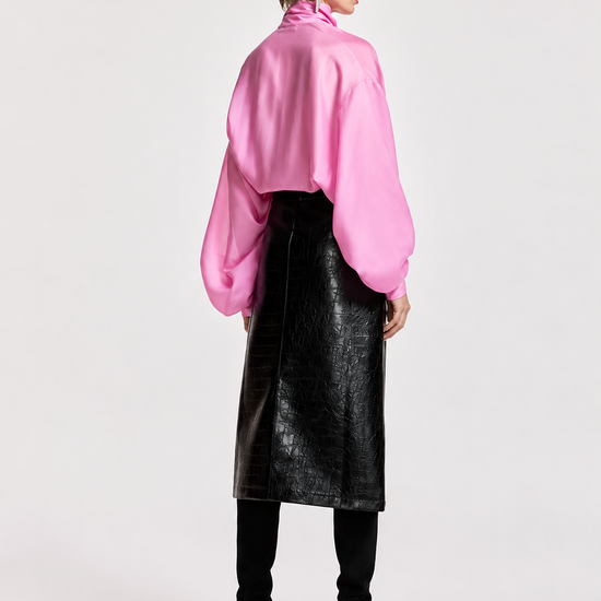 A model is shown from the back wearing a vibrant pink long-sleeve blouse with voluminous balloon sleeves, a black faux-leather midi skirt, black leggings, and black high-heeled shoes, set against a plain light gray studio background.