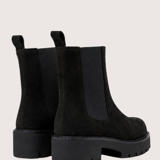 A pair of black Chelsea boots with chunky lug soles and elastic side panels, shown from a rear three-quarter angle on a light beige background.
