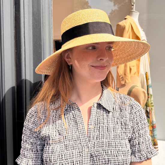 Woman wearing a wide-brimmed hat with a black band, standing in front of a store window.
