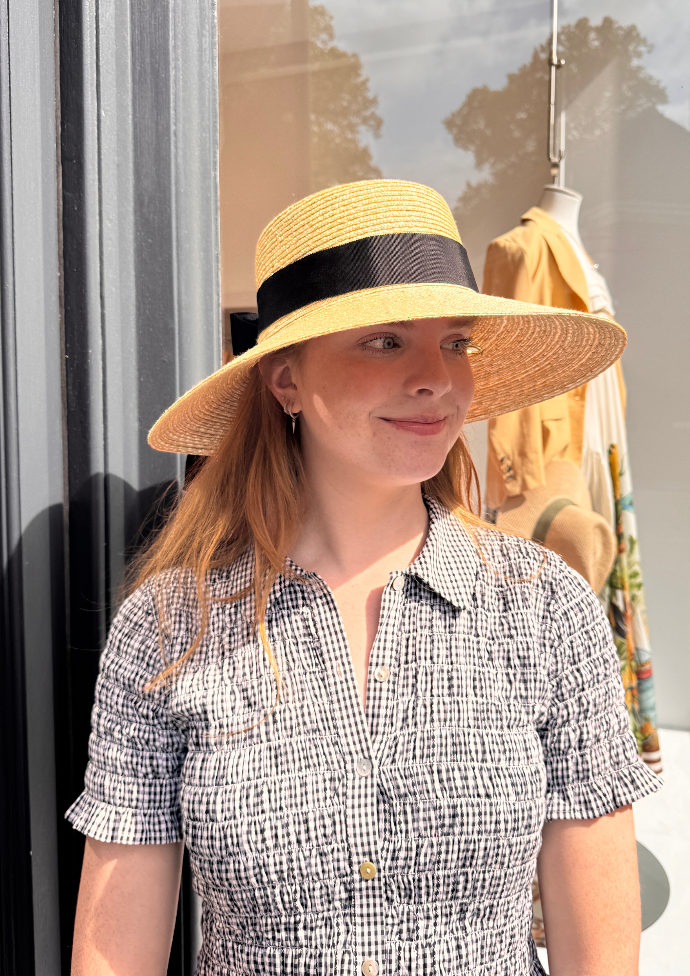 Woman wearing a wide-brimmed hat with a black band, standing in front of a store window.
