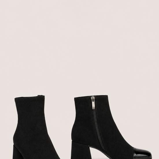 A pair of black ankle boots with a block heel and glossy patent leather toe caps, shown against a solid light beige background.