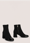 A pair of black ankle boots with a block heel and glossy patent leather toe caps, shown against a solid light beige background.