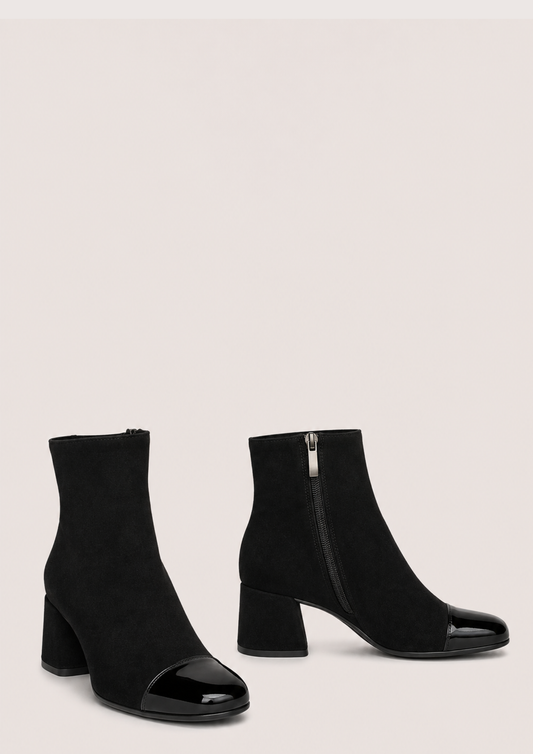 A pair of black ankle boots with a block heel and glossy patent leather toe caps, shown against a solid light beige background.
