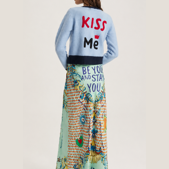 A model is shown from behind wearing a light blue cardigan with 'KISS Me' printed on the back in red and black, paired with a long, colorful patterned maxi skirt featuring 'BEYOND AND STAY YOU' and 'KISS ME' text.