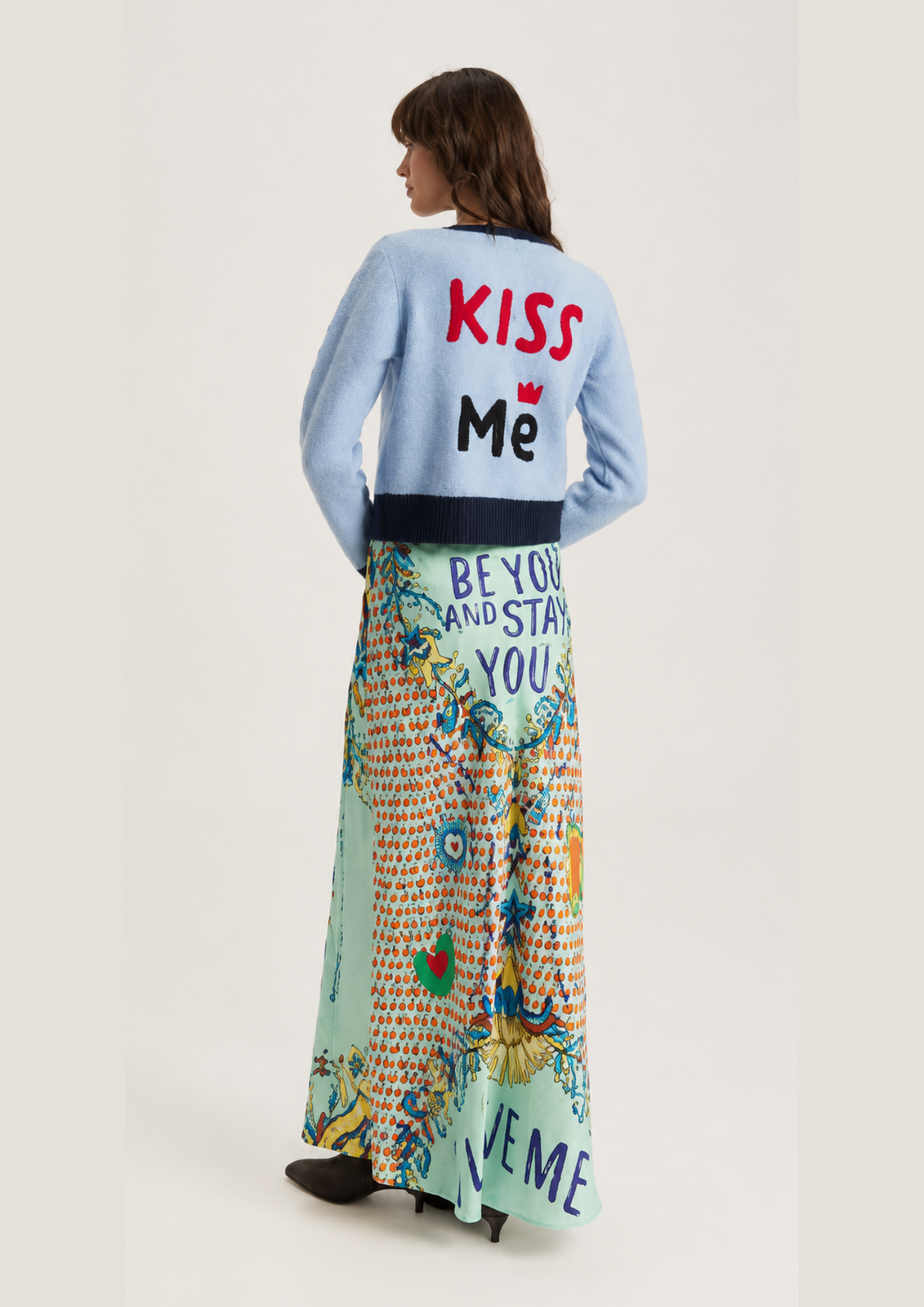 A model is shown from behind wearing a light blue cardigan with 'KISS Me' printed on the back in red and black, paired with a long, colorful patterned maxi skirt featuring 'BEYOND AND STAY YOU' and 'KISS ME' text.