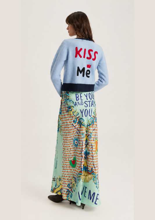 A model is shown from behind wearing a light blue cardigan with 'KISS Me' printed on the back in red and black, paired with a long, colorful patterned maxi skirt featuring 'BEYOND AND STAY YOU' and 'KISS ME' text.