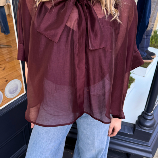 Women wearing burgundy sheer blouse with tie neck and blue jeans, close up image.