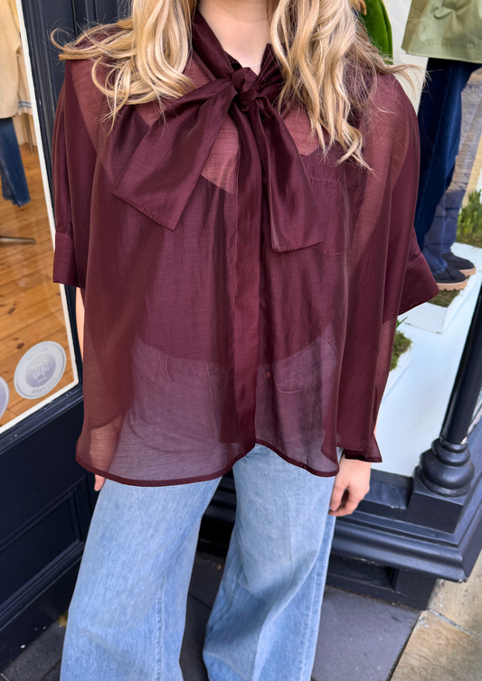 Women wearing burgundy sheer blouse with tie neck and blue jeans, close up image.