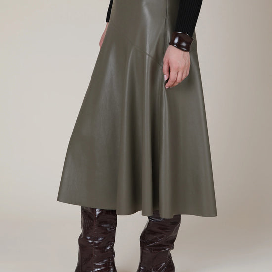 A model is shown from the waist down wearing a high-waisted, A-line midi skirt in a smooth, matte gray faux leather. The skirt is styled with dark brown pointed-toe heeled boots and a black long-sleeve top against a neutral beige studio background.