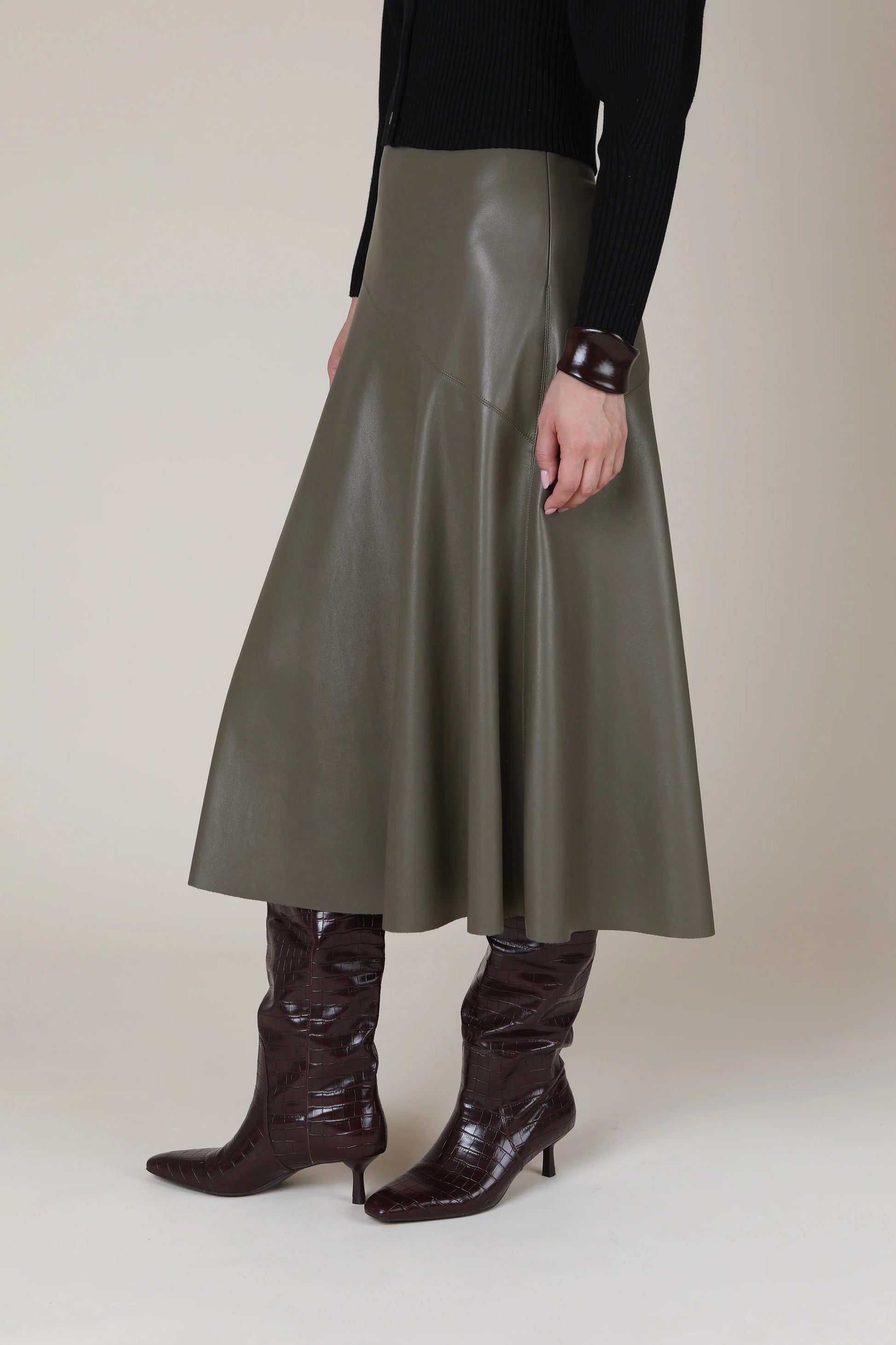 A model is shown from the waist down wearing a high-waisted, A-line midi skirt in a smooth, matte gray faux leather. The skirt is styled with dark brown pointed-toe heeled boots and a black long-sleeve top against a neutral beige studio background.