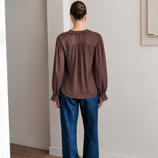 A woman wearing a brown long-sleeve blouse with gathered ruffles at the collar and cuffs, paired with blue jeans, sitting on a light gray upholstered chair.