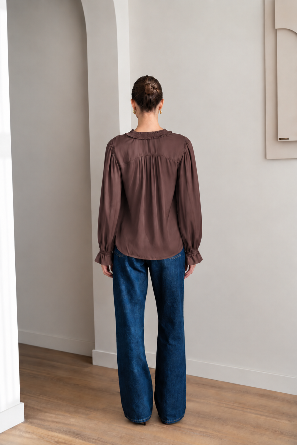 A woman wearing a brown long-sleeve blouse with gathered ruffles at the collar and cuffs, paired with blue jeans, sitting on a light gray upholstered chair.