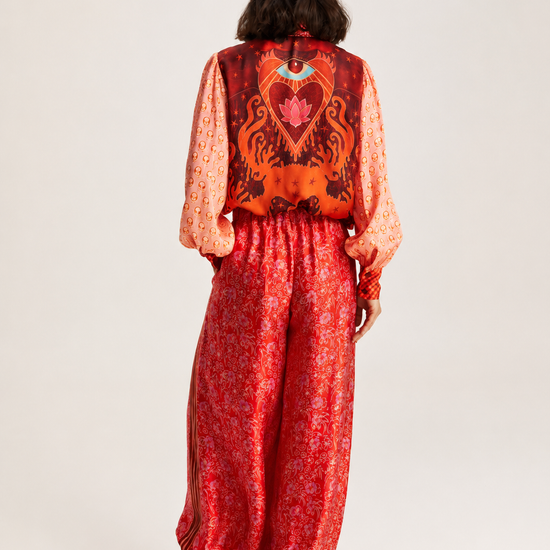 A model viewed from the back wearing a vibrant red and orange patterned jumpsuit with a large heart design on the back, paired with black ankle boots, against a plain light gray studio background.